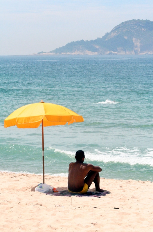 Man sitting on sand under a yellow beach umbrella.