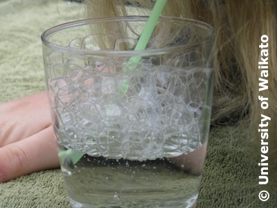 Person with a green straw blowing bubbles in a glass.