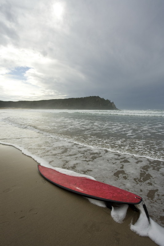 A red Flax (harakeke) surfboard on a beach by waters edge.