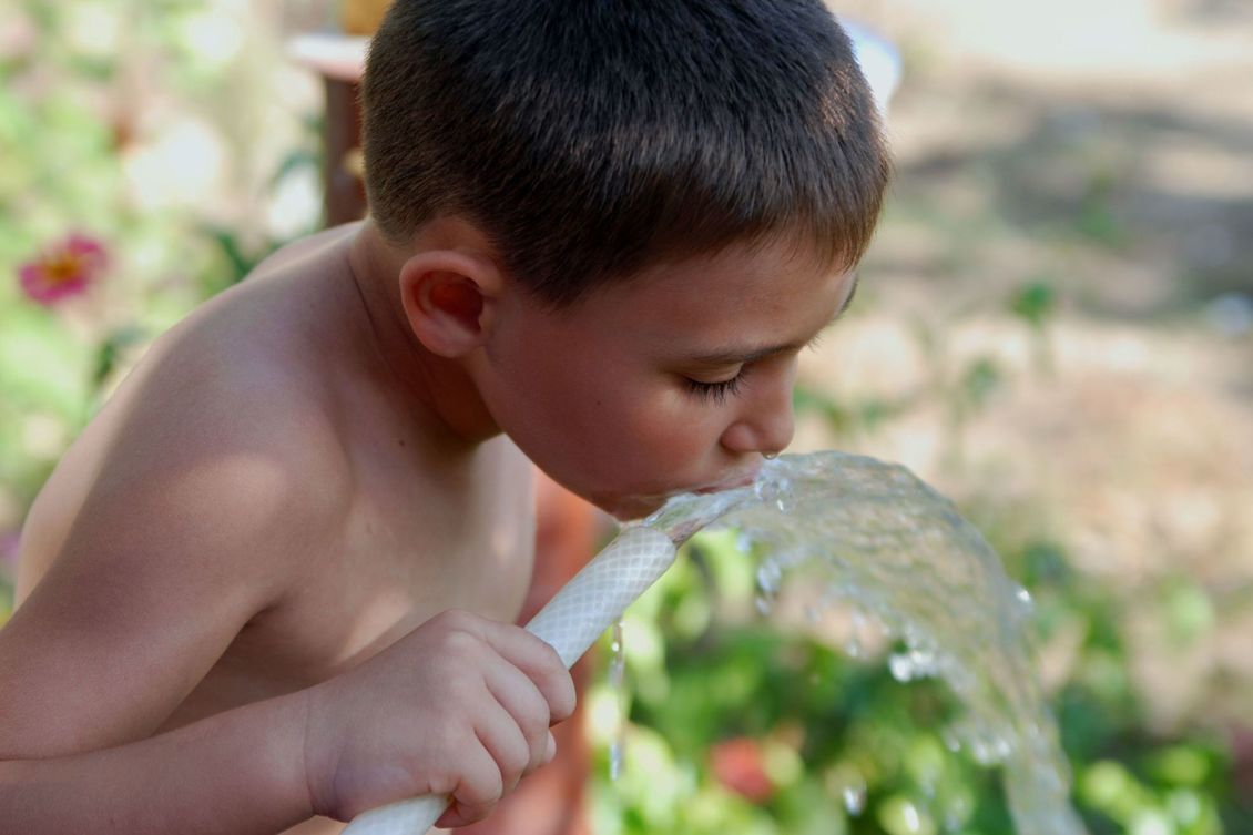 Young boy drinking water from a garden hose.