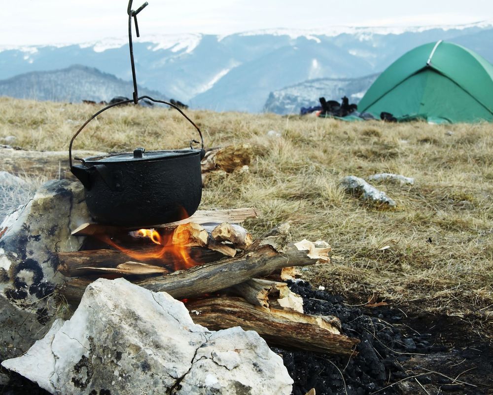 Pot on a camp fire with green tent and mountains in background